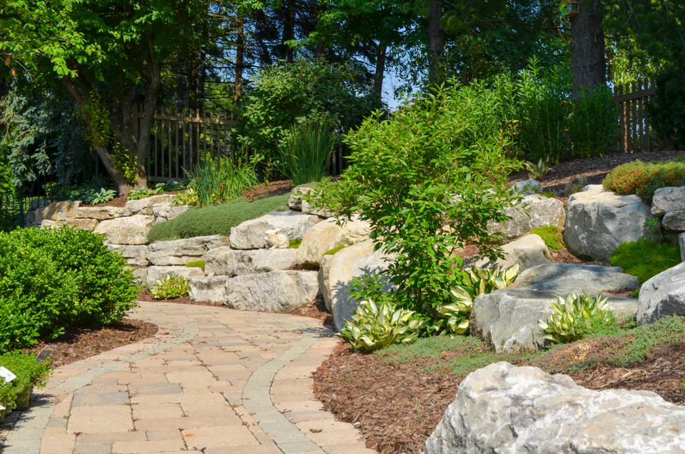 Paver garden walk with limestone boulders, evergreen shrubs, mulch beds, and wooded privacy edge.
